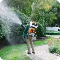 A Mainely Grass technician applying mosquito and tick control treatment in a well-maintained yard, with a branded service van in the background.